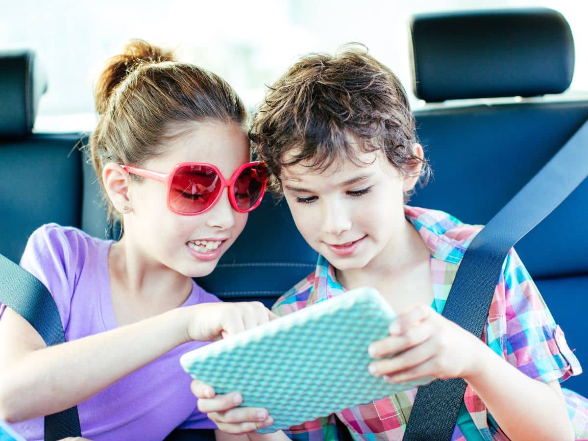 tween boy and girl smiling and watching movie on device in backseat of car