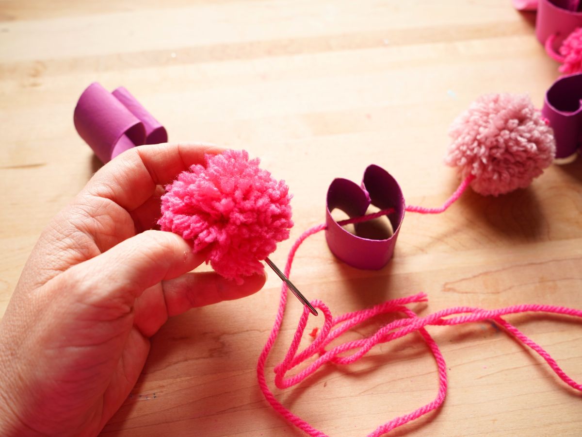 pink pom pom with embroidery needle poking through center and onto yarn garland